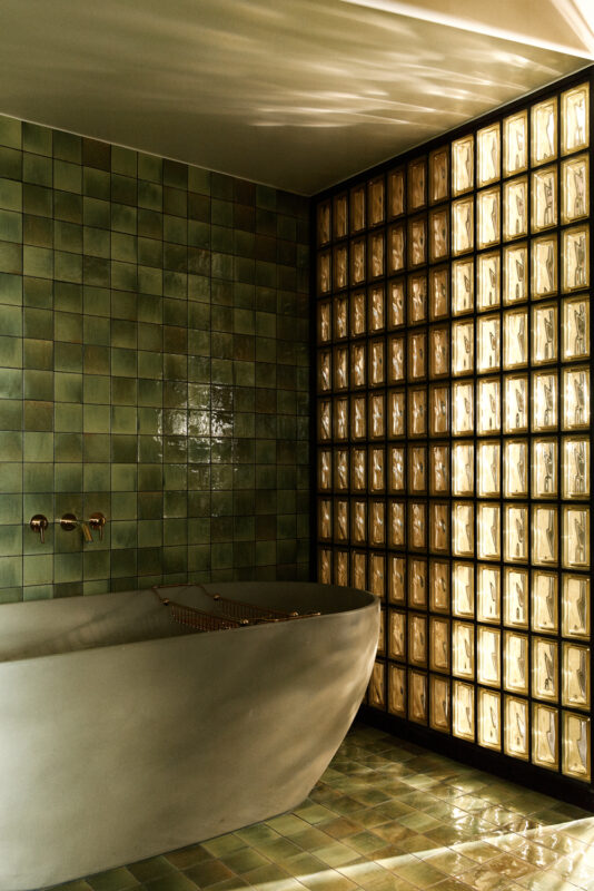 Luxurious bathroom featuring a curved bathtub, green tiled walls, and a glass block wall that reflects warm light.
