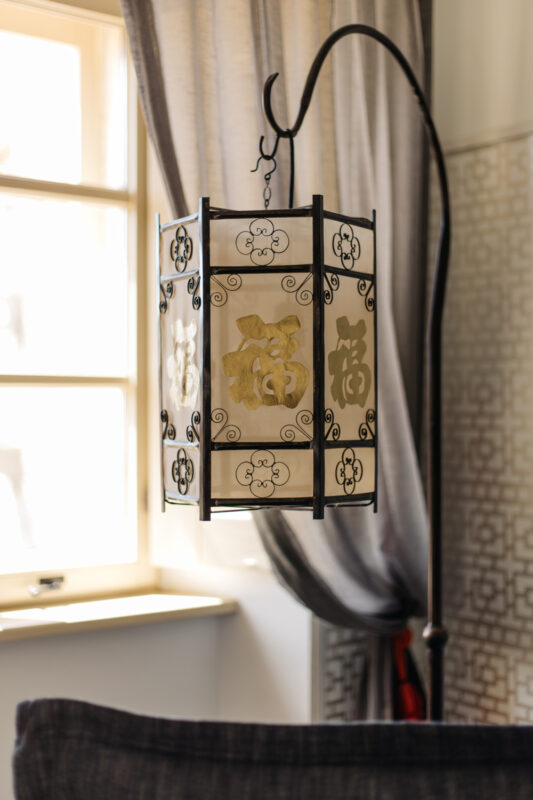 Chinese lantern with decorative patterns, hanging from a curved stand, in a well-lit room with large windows.
