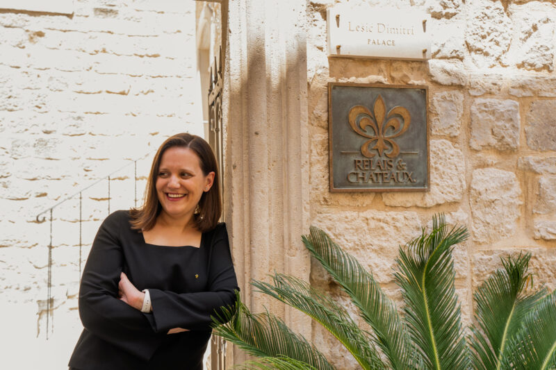 Woman smiling and leaning against a wall near the Lešić Dimitri Palace sign, with a palm plant in the foreground.