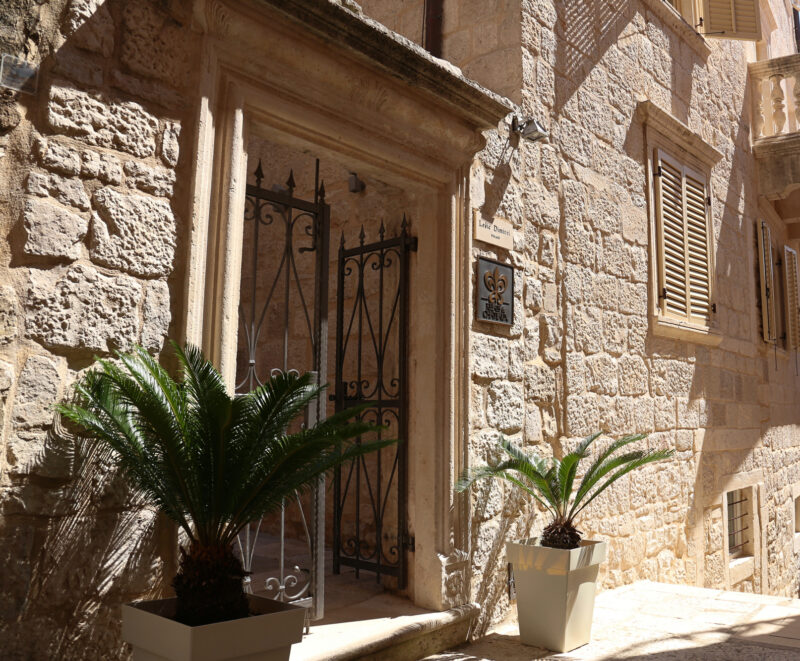 Stone building entrance with wrought iron gate, flanked by potted palm trees, in a narrow street of Lešić Dimitri Palace.