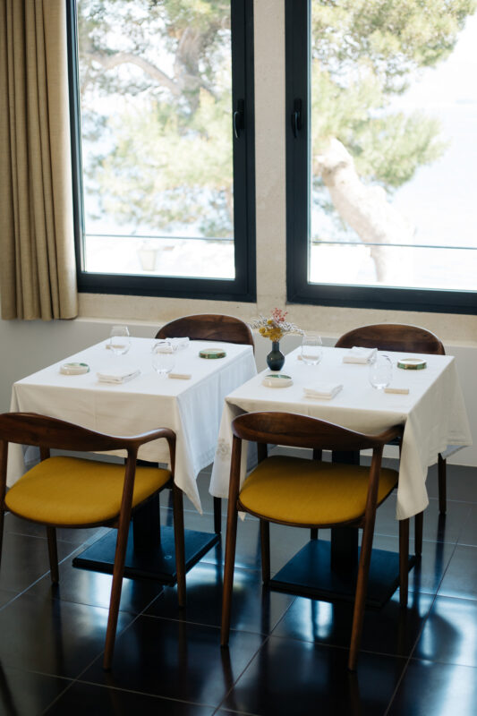 Elegant dining setup with two tables, white tablecloths, and glassware, overlooking a scenic view through large windows.