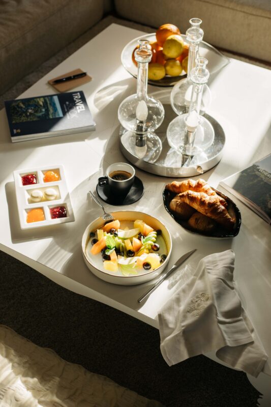 Breakfast spread on a coffee table featuring fruit, yogurt, croissant, and coffee, with books and decor in the background.