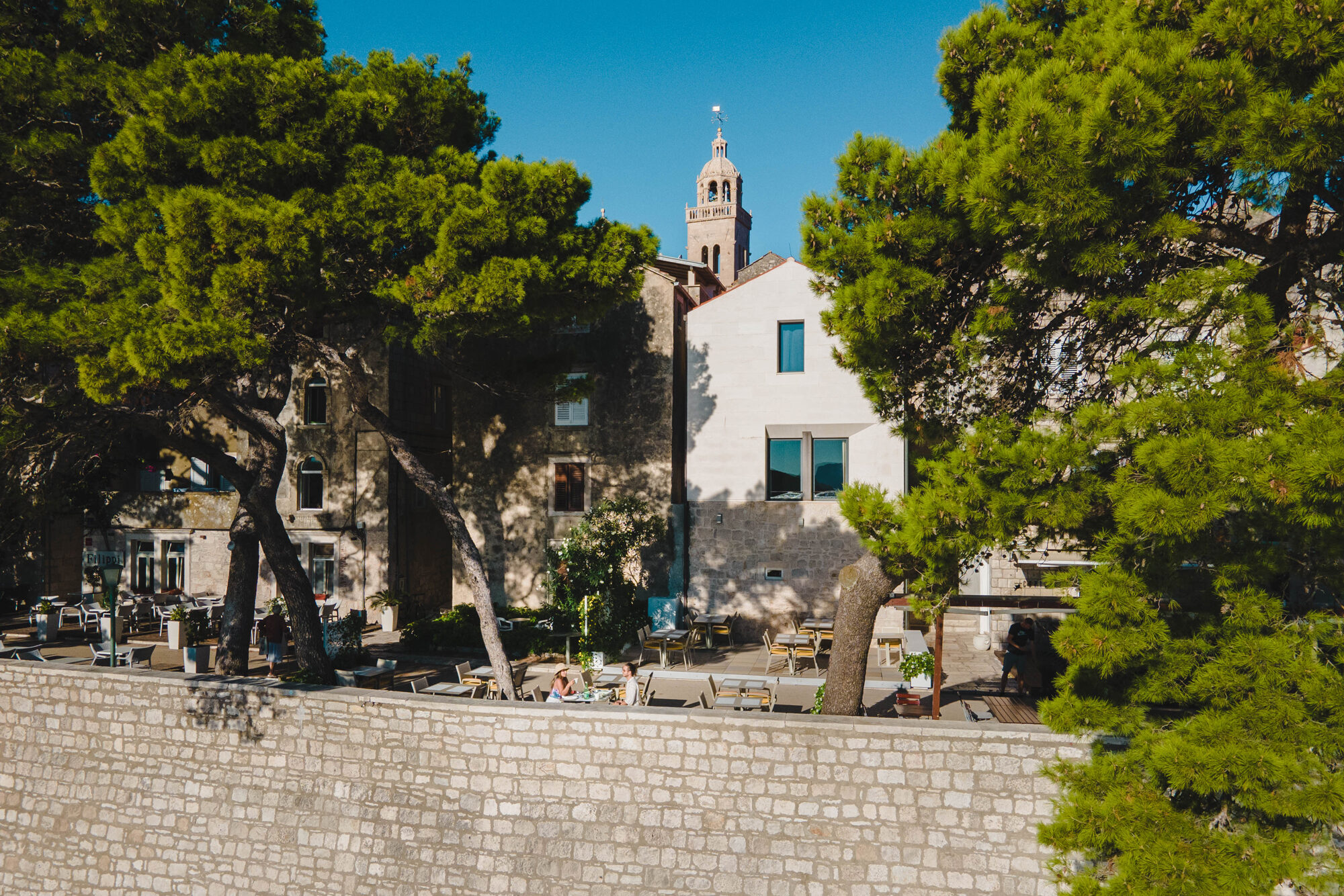 Historic buildings with a stone wall and trees, featuring a church tower in the background, at Lešić Dimitri Palace.