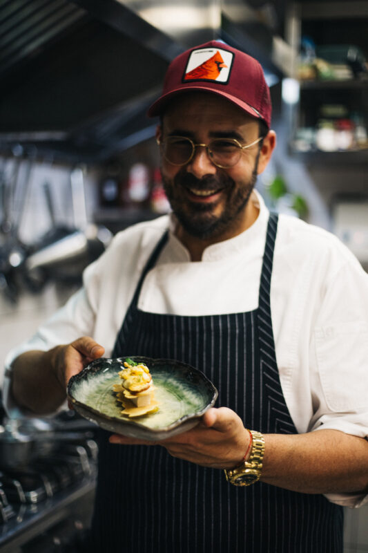 Chef Marko Gajski in a kitchen, smiling while presenting a dish on a decorative plate.