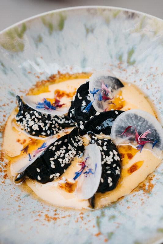Black gyoza arranged in a circular pattern, garnished with edible flowers and sesame seeds, served in a creamy sauce.