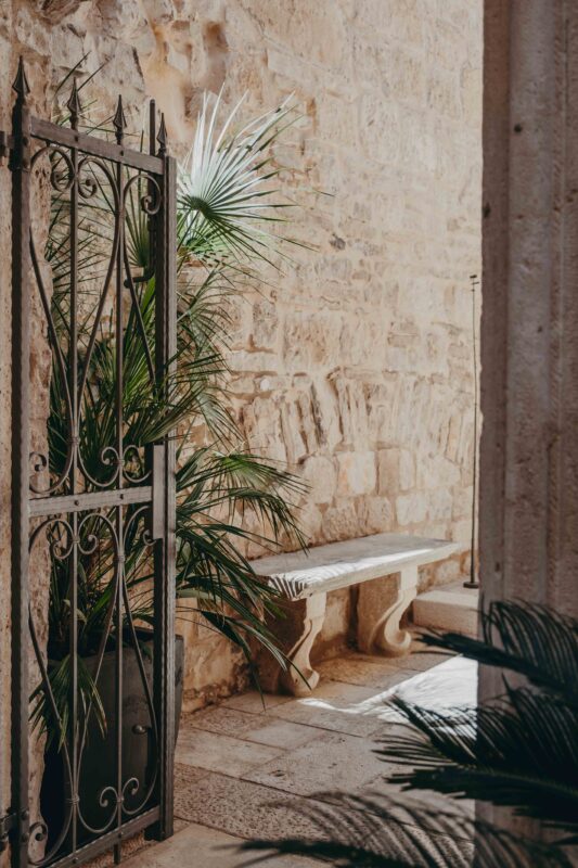 Stone wall with a wrought iron gate, a bench, and tropical plants in the entrance of Lešić Dimitri Palace.