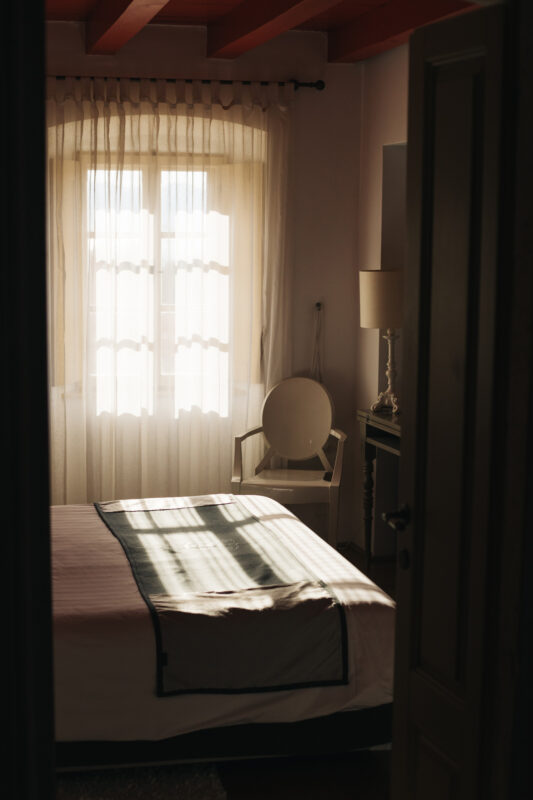 Brightly lit bedroom with a bed, chair, and window, featuring soft curtains and warm wooden beams.