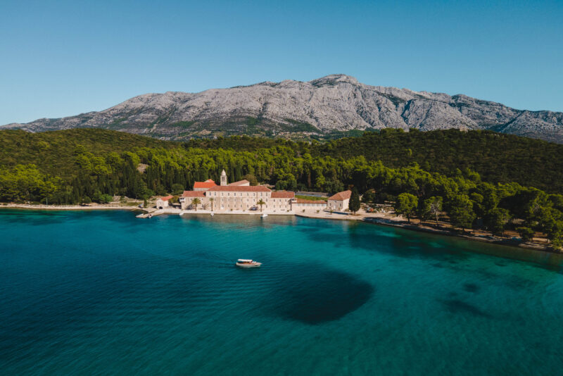 Aerial view of Lešić Dimitri Palace by the turquoise sea, with a boat anchored nearby and mountains in the background.