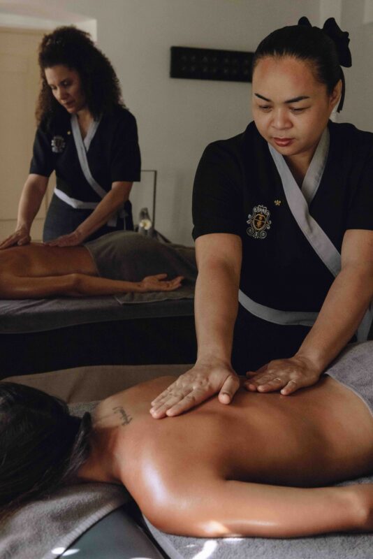 Two therapists performing a massage in a serene spa setting, with one applying pressure to a client's back.