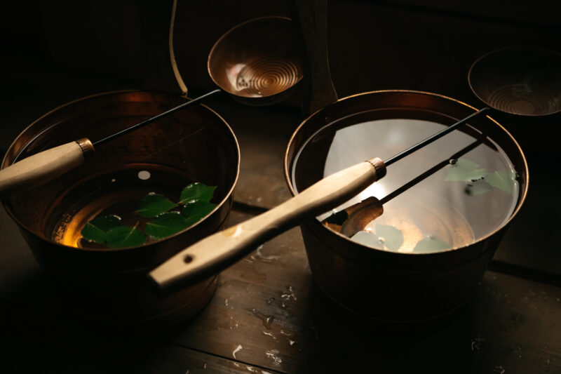 Two wooden ladles rest on the edge of copper buckets filled with water and floating green leaves, set in a sauna environment.