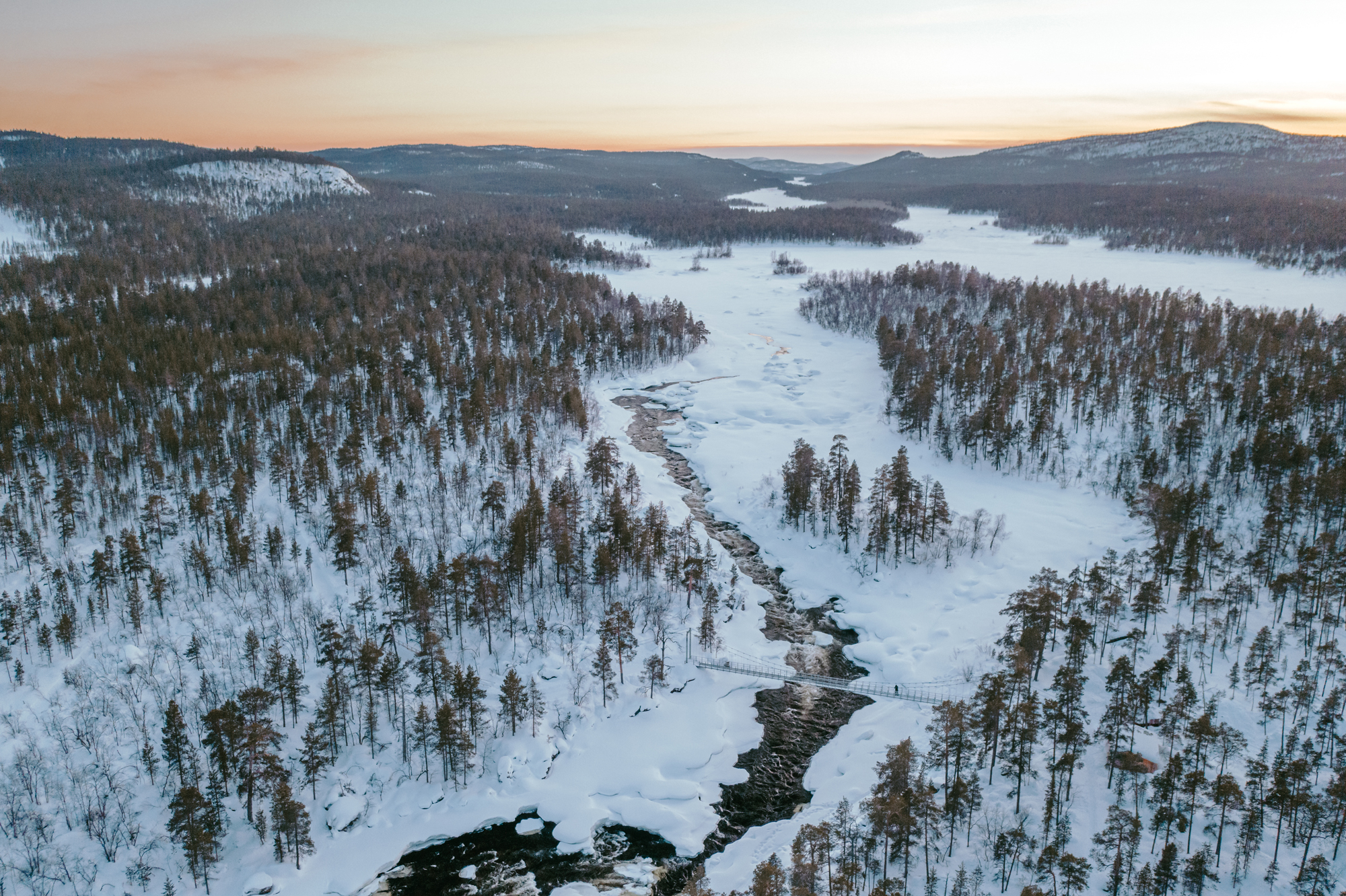Snow-covered landscape with a winding river, surrounded by dense forests and mountains under a soft sunset sky.