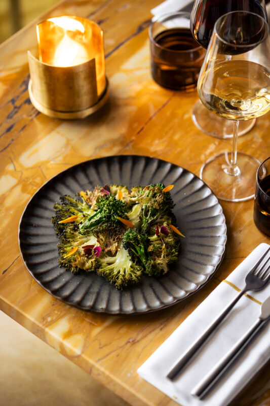 Plate of roasted greens garnished with colorful microgreens, set on a textured black plate, with wine glasses and a candle...