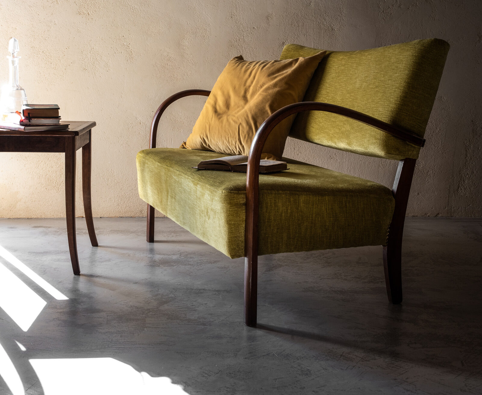 A green upholstered loveseat with a yellow cushion beside a wooden side table and books, illuminated by natural light.