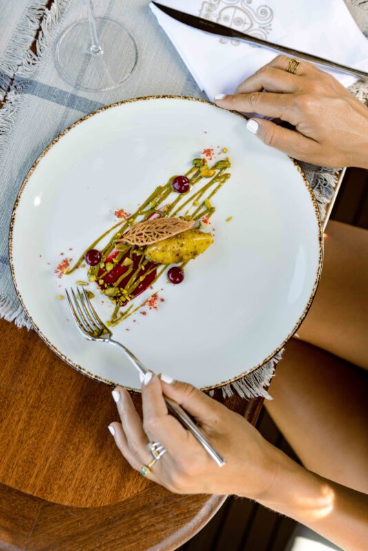 Elegant dish on a white plate with colorful garnishes, held by a person with manicured nails, on a wooden table.