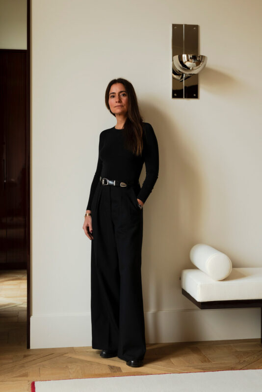 Portrait of Marika Dru standing against a light wall, wearing a black long-sleeve top and high-waisted trousers.