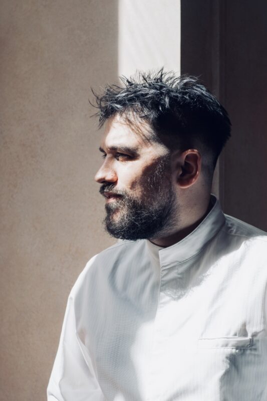 Portrait of Chef Thomas Rossi, featuring him in profile with a light source illuminating his face against a textured backg...
