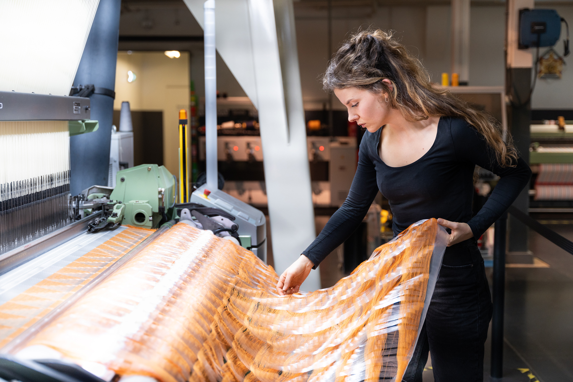 Young woman examining a large piece of fabric at a textile loom in a workshop setting.
