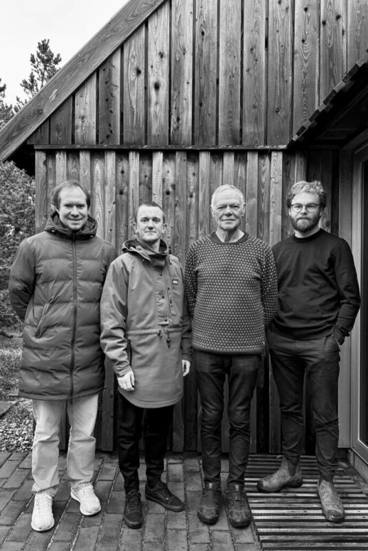 Four men standing in front of a wooden building, dressed casually, with trees in the background.
