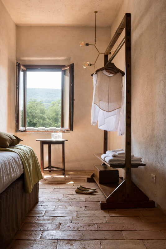 Bright, minimalist room featuring a bed, wooden clothing rack with garments, a small table, and a window with a scenic view.