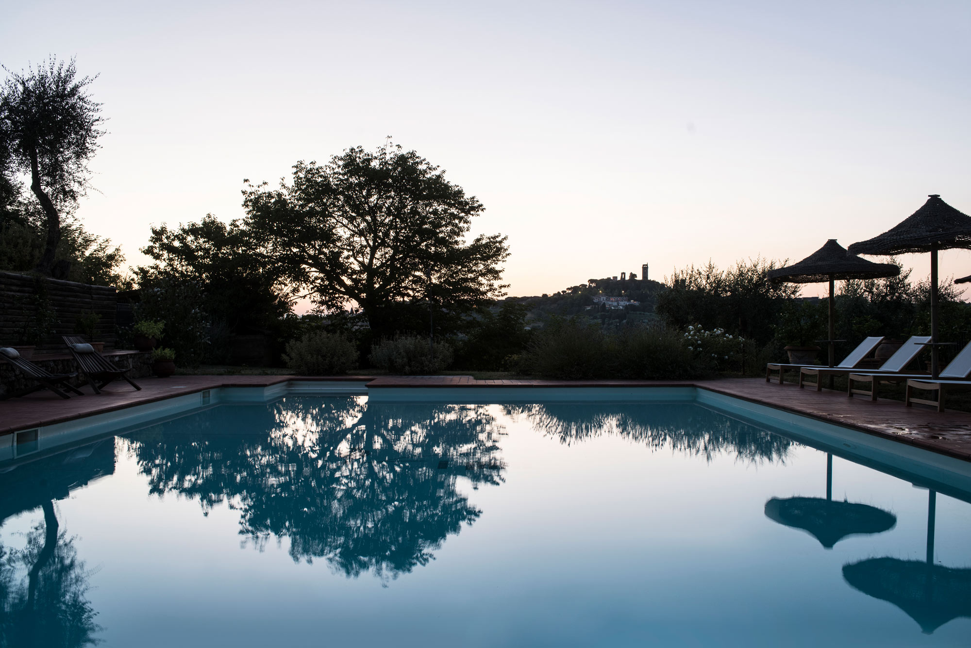 Tranquil saltwater pool at dusk, reflecting trees and distant hills with silhouettes of lounge chairs and umbrellas.