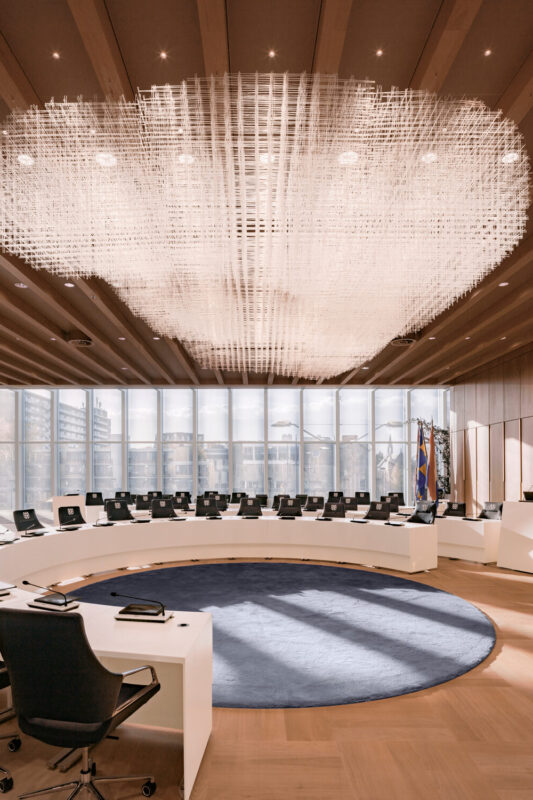 Modern council chamber in Tilburg featuring a circular seating arrangement, large chandelier, and floor-to-ceiling windows.