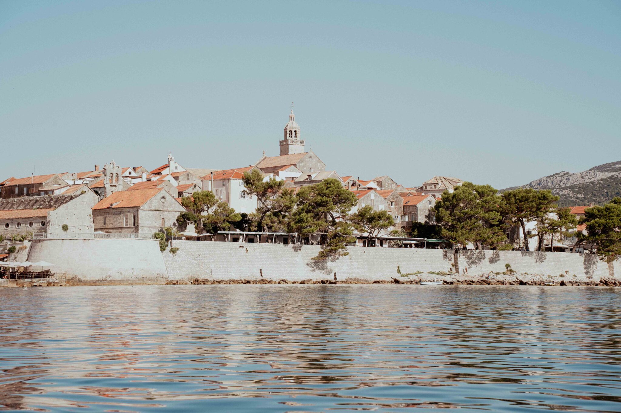 Coastal view of Lešić Dimitri Palace with red-roofed buildings and trees reflected in calm water.
