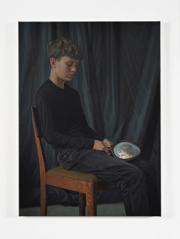 A boy sits on a wooden chair, holding a small mirror reflecting a child's face, against a dark curtain backdrop.