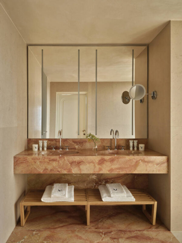 Elegant bathroom featuring a marble sink with two faucets, wooden benches, and a large mirror, in the VO Corner Junior Suite.