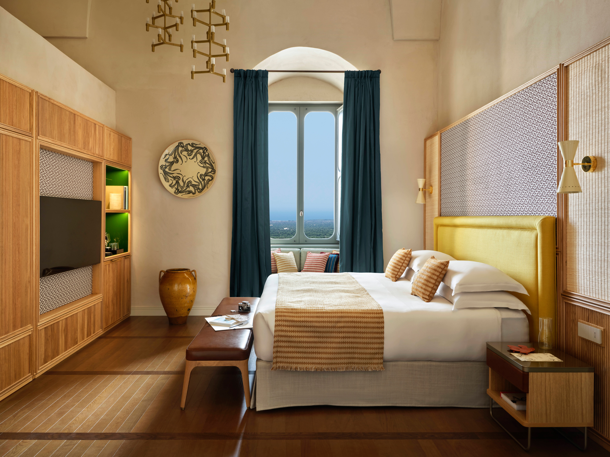 Elegant hotel room featuring a yellow headboard, wooden furniture, and large window with a view of Ostuni.
