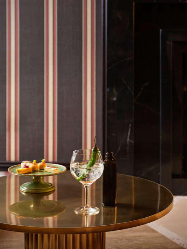 A glass of tonic water with a green chili sits on a round gold table beside a plate of colorful appetizers.