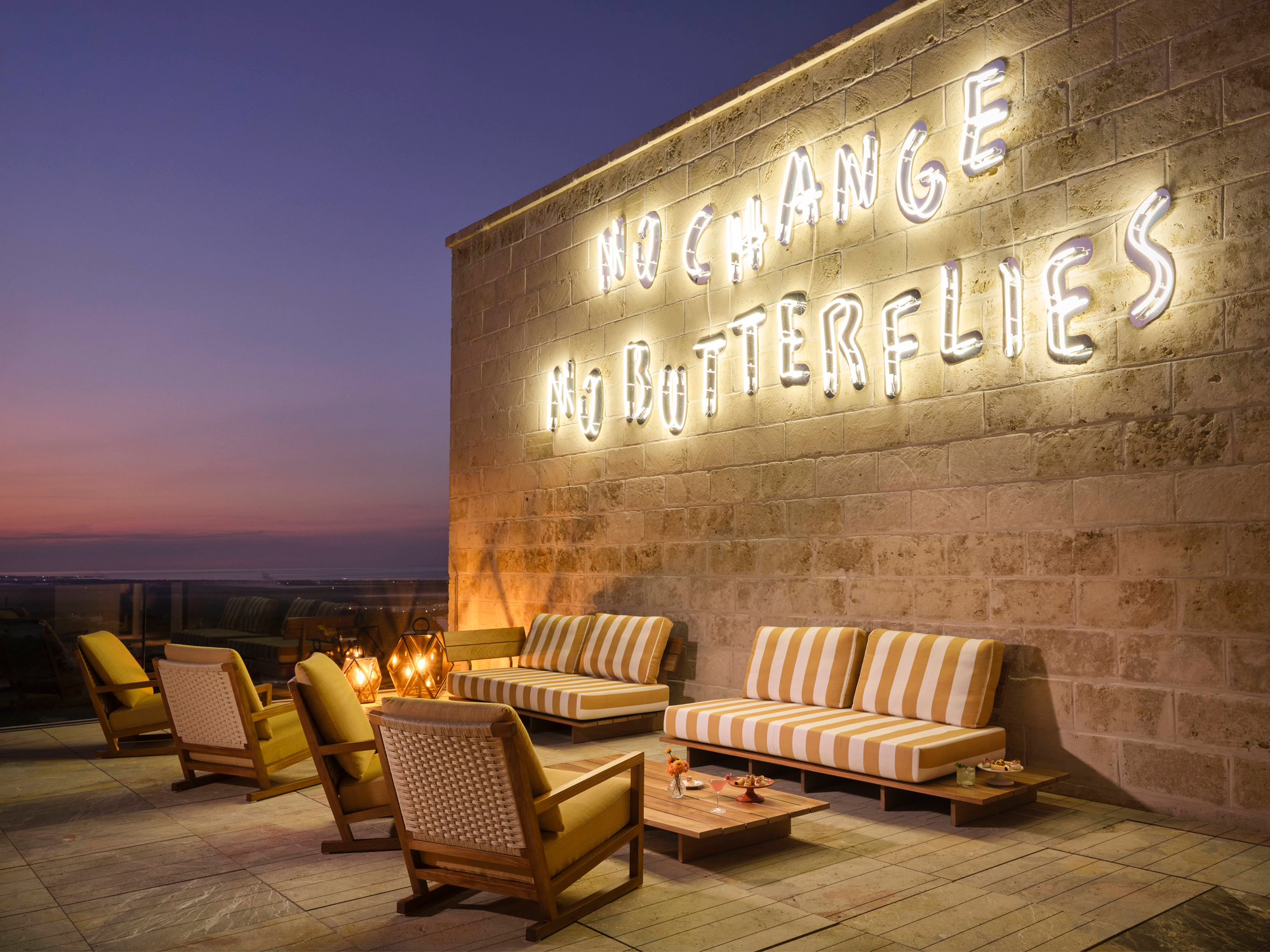 Cozy outdoor seating area at Infinity Bar with a neon sign reading "NO CHANGE NO BUTTERFLIES" at sunset.