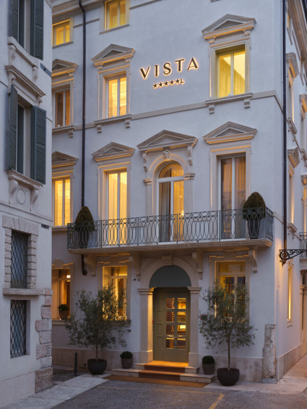 Elegant hotel facade with "VISTA" signage, warm lights glowing from windows, and potted plants at the entrance.