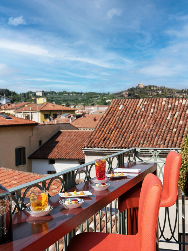 Colorful cocktails on a balcony overlooking Verona's rooftops and distant hills under a blue sky.