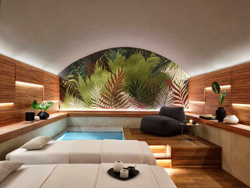 Spa room featuring two massage tables, a small plunge pool, and a tropical mural, with wooden accents and plants.