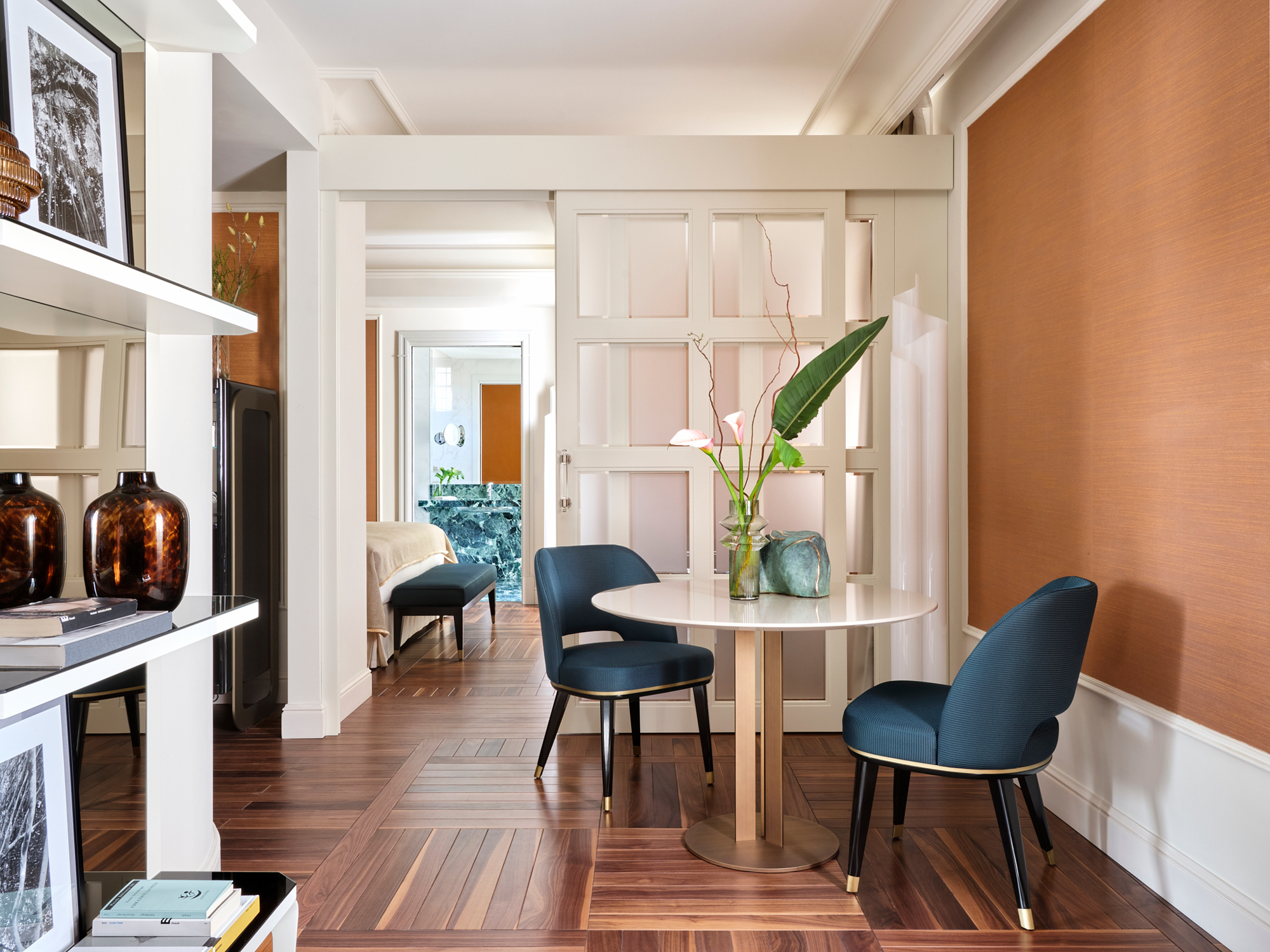 Modern interior of a room featuring a round table with two blue chairs, a vase with flowers, and wooden flooring.