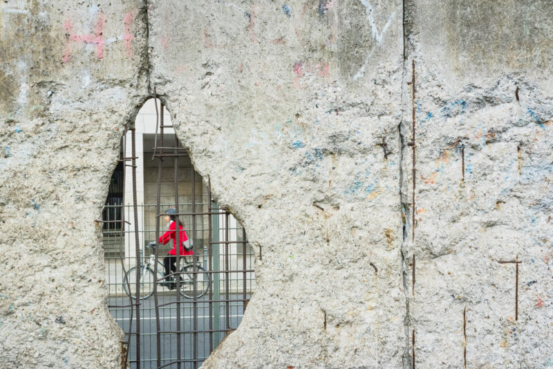 A cyclist in a red jacket rides past a cracked concrete wall with a large hole revealing the street beyond.