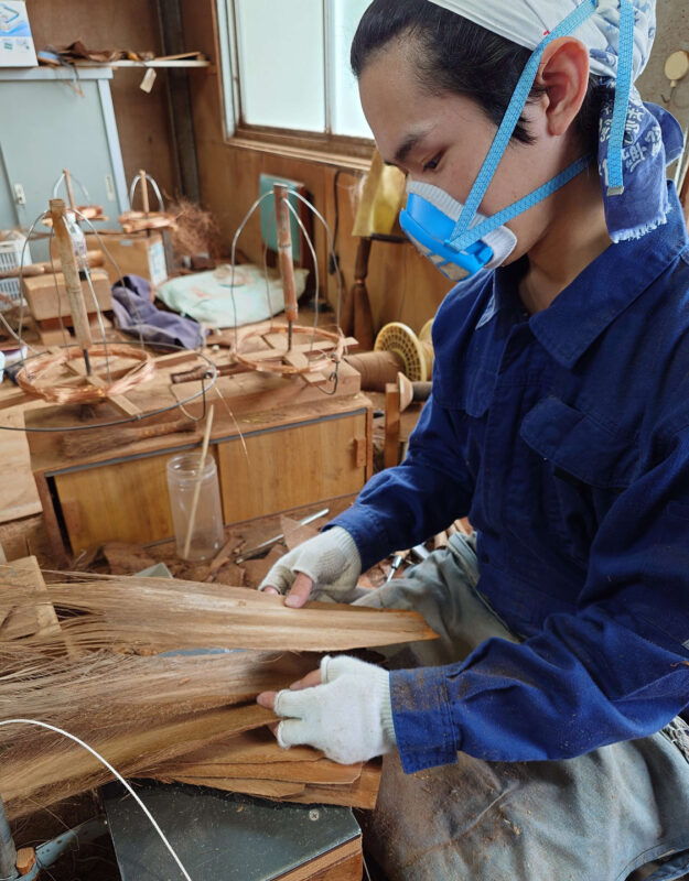 Young man in blue work clothes and mask, crafting wood in a workshop filled with tools and wooden materials.