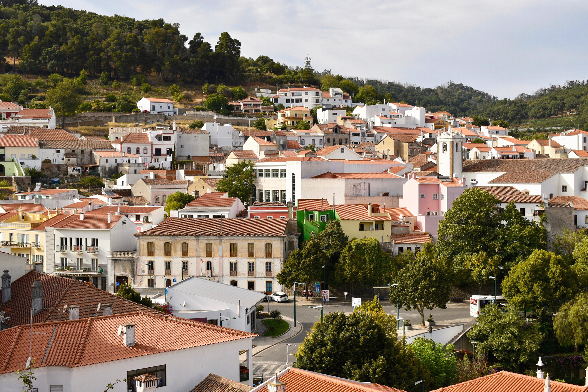 Colorful buildings with red-tiled roofs nestled in a hilly landscape, featuring trees and a church tower in the background.