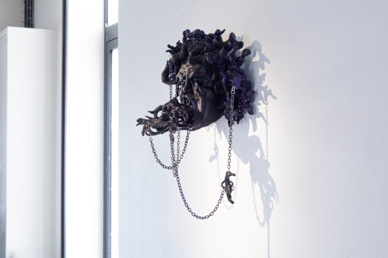 Medusa self-portrait sculpture by Adèle Vivet, featuring dark, textured elements and chains against a white wall.