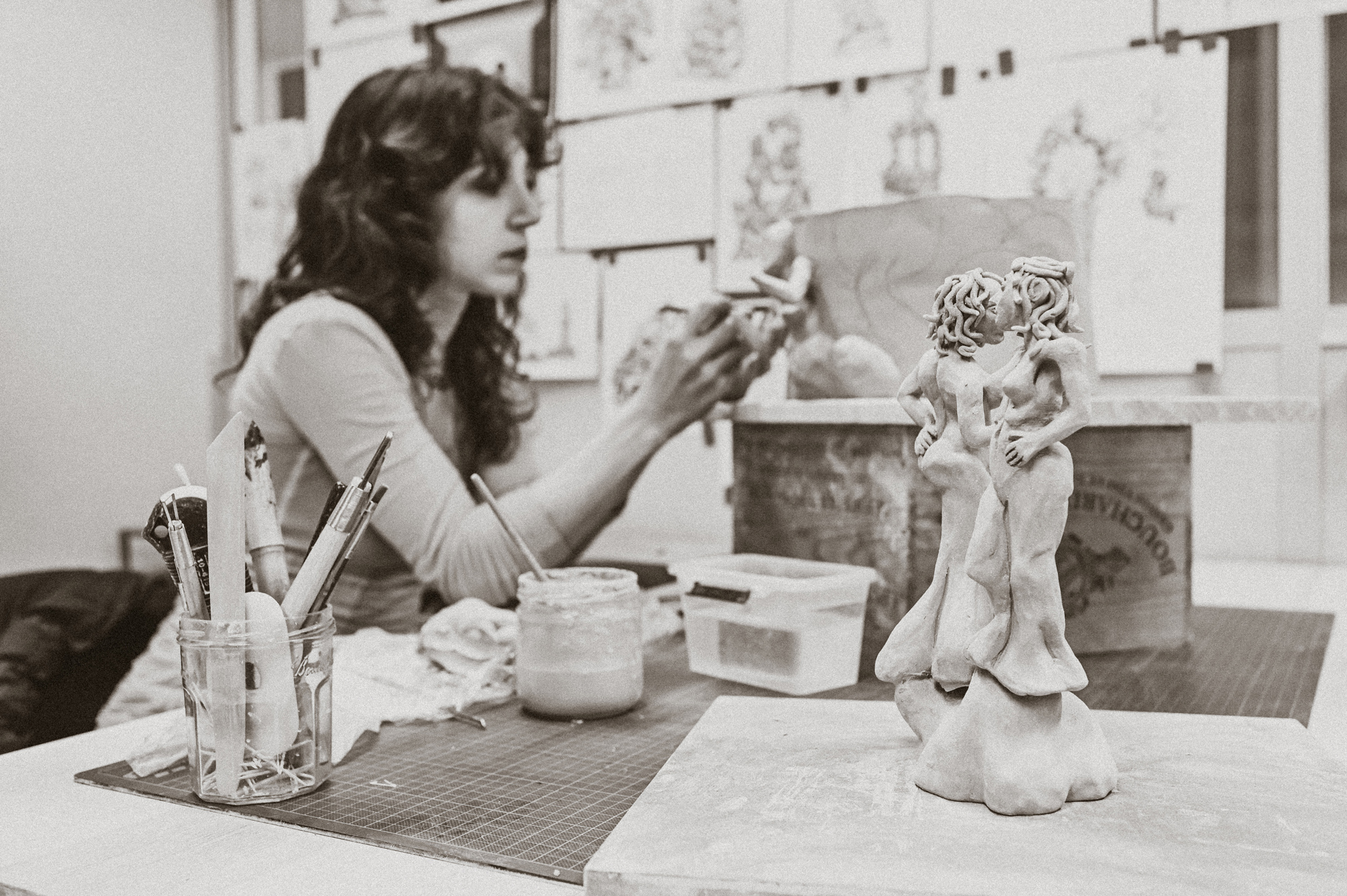 Artist Adèle Vivet sculpting a clay figure in a studio, with art supplies and sketches in the background.