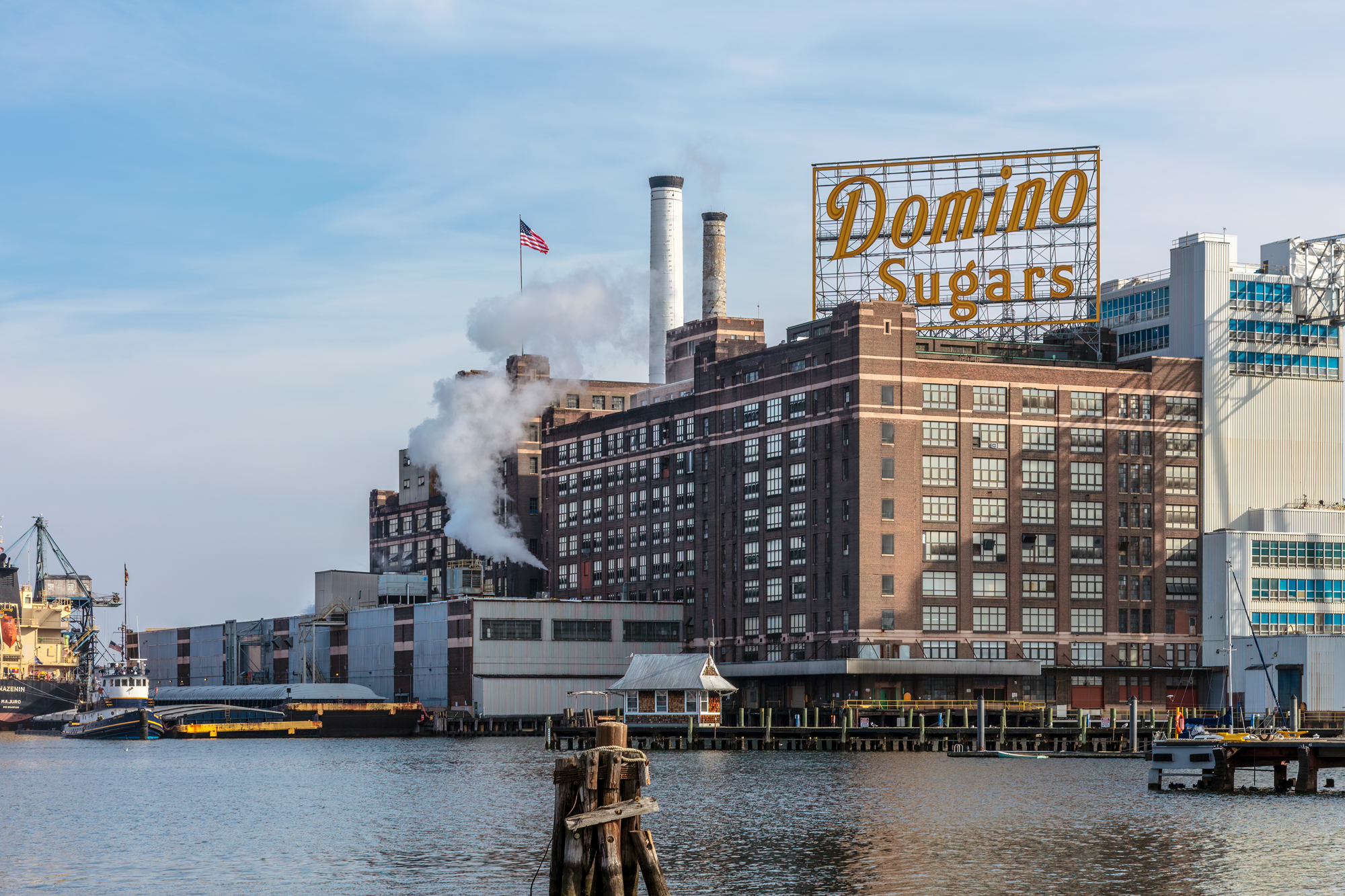 Domino Sugars refinery in Baltimore, featuring a large sign and smoke rising from the building, with water in the foreground.
