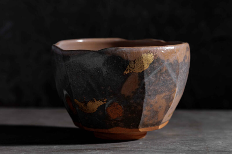 Ceramic bowl with a textured surface and gold leaf accents, set against a dark background.