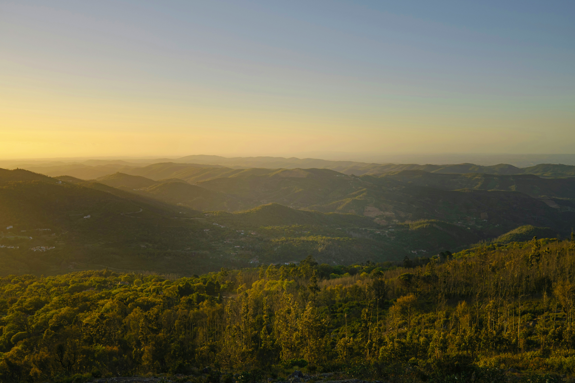 Sunset over rolling hills and valleys, with lush greenery and distant mountains under a clear sky.