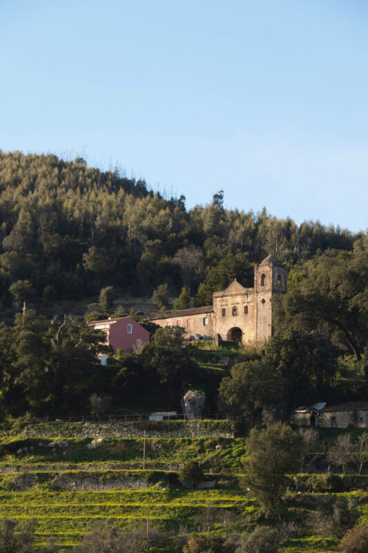 Historic building on a hillside, surrounded by trees and greenery, with a clear blue sky above.