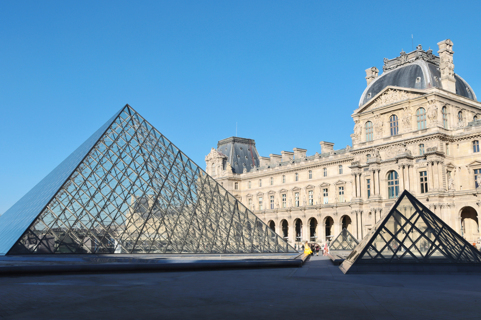 Glass pyramids of the Louvre Museum against a clear blue sky, with the historic building in the background.