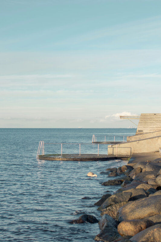 Calm shoreline with rocks, a wooden platform extending into the water, and a clear blue sky.
