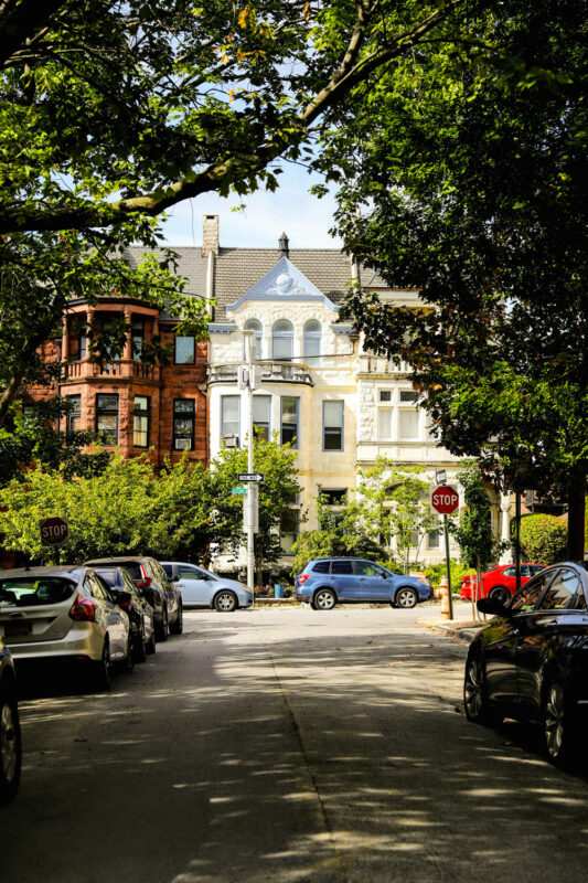 Row of residential buildings with trees lining the street, featuring a mix of parked cars and stop signs.