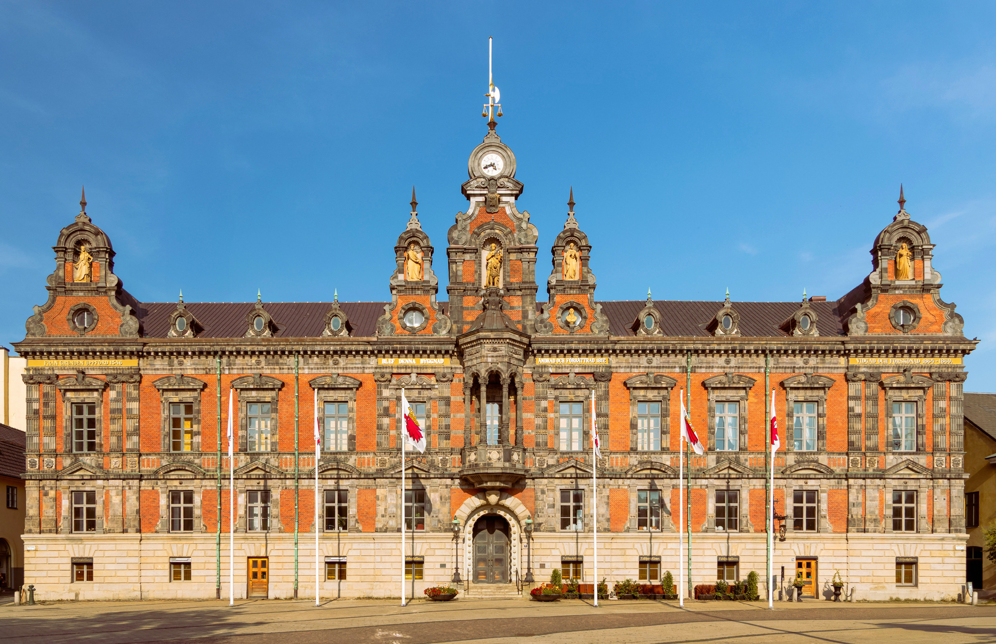 Historic building with ornate architecture, featuring a clock tower and multiple flags in front against a clear blue sky.