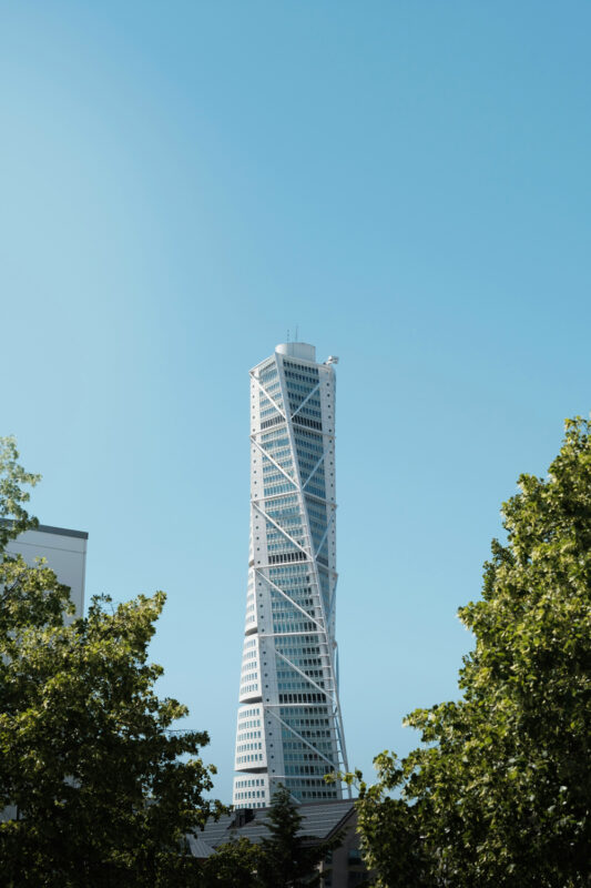 Tall white twisted skyscraper with external diagonal braces, rising above trees under a clear blue sky.