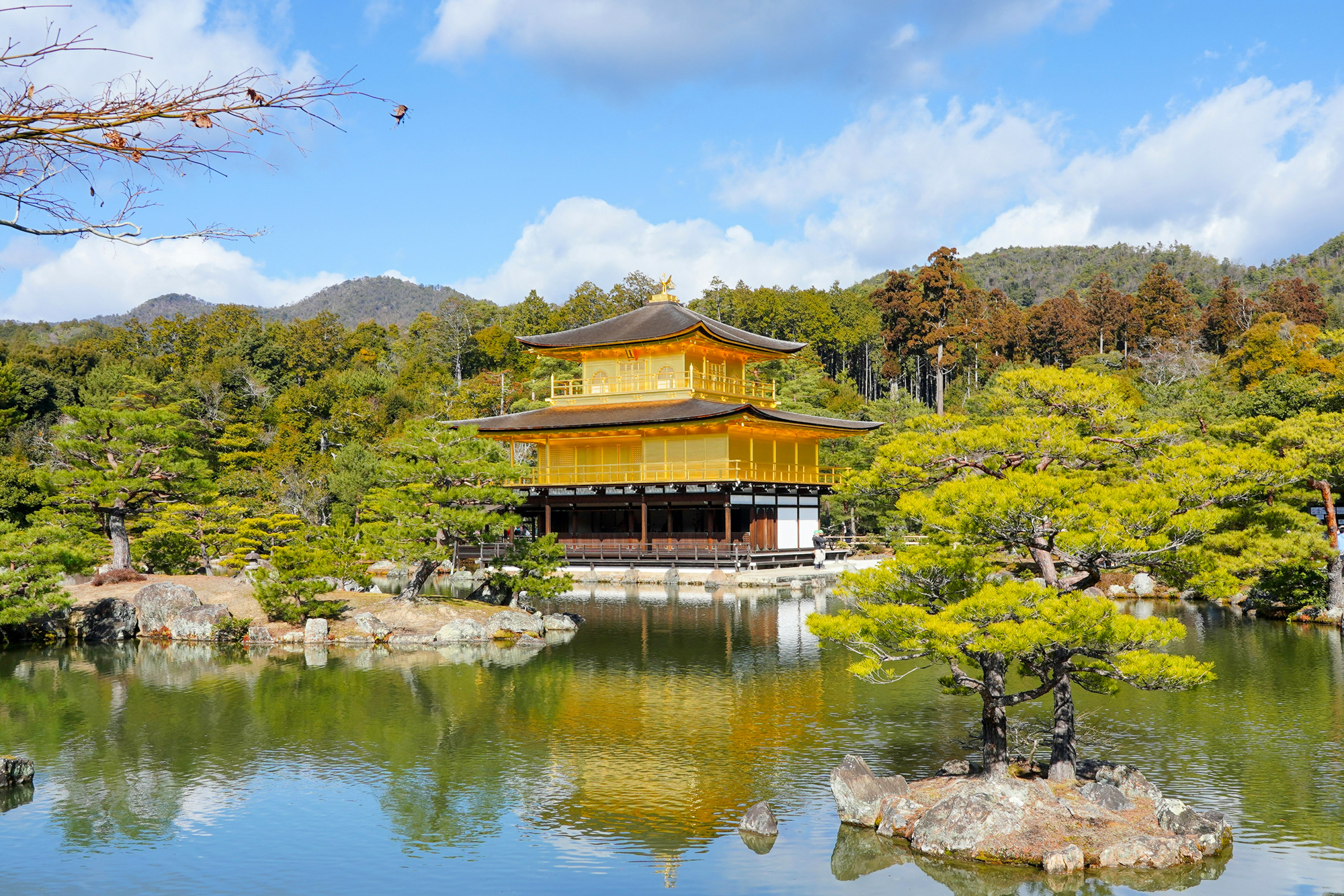 Golden pavilion surrounded by trees and water, reflecting the sky and mountains in the background.